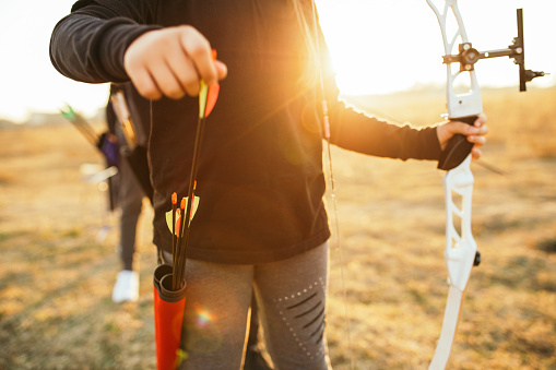 Child girl pulling out arrow from her quiver on outdoors archery training at sunset and holding bow in other hand