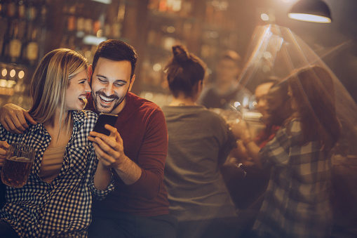 Young happy couple reading a text message on smart phone while spending their night in a bar. There are people in the background.