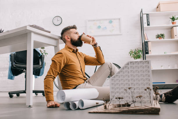 istock bearded adult male architect drinking coffee to go and working on construction project with blueprints in office 1070530686