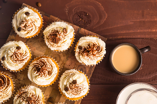 Tiramisu cupcakes decorated with cocoa powder and coffee grains on a wooden board next to jugs of milk, cream and a cup of coffee. Top view, flat lay