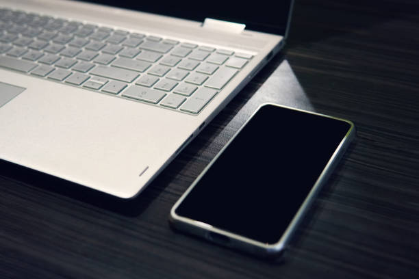 Smart phone with copy space screen and laptop on wooden table background. Empty black screen of a mobile left on the table near the computer. Use of digital gadgets at work. Phone display mockup