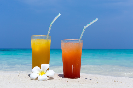 Two Cocktails in glass on white sandy beach decorated plumeria blossom with sea background. Travel card with copy space, nobody