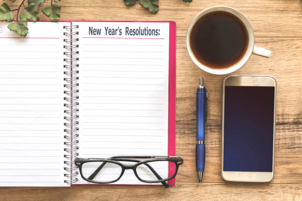Office desk with "New Year's Resolutions" note and cell phone. Wooden office desk with items surrounding a "New Year's Resolutions" note pad. Group of objects includes: glasses, note pad, cup of coffee, pen and cell phone. No people. 2019 new year resolution stock pictures, royalty-free photos & images