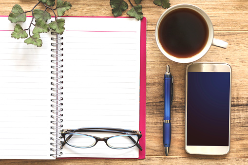 Wooden office desk with items surrounding a blank note pad.  Group of objects includes: glasses, note pad, cup of coffee, pen and cell phone. No people.