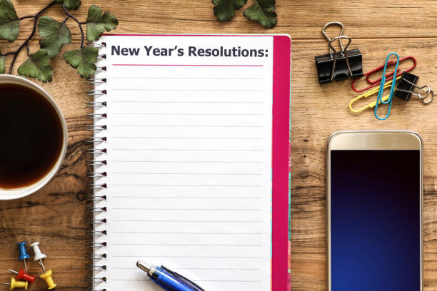 Office desk with "New Year's Resolutions" note and cell phone. Wooden office desk with items surrounding a "New Year's Resolutions" note pad. Group of objects includes: note pad, pen, fasteners, cup of coffee and cell phone. No people. 2019 new year resolution stock pictures, royalty-free photos & images