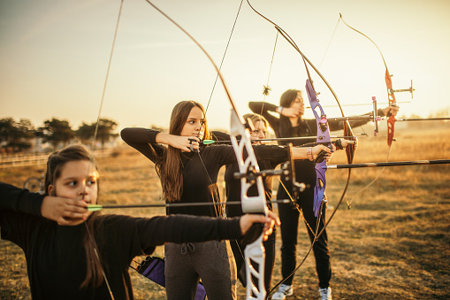 Group of females on outdoors archery training at sunset. They are all aiming with bow and arrow. The group includes mid adult women, children, teenagers