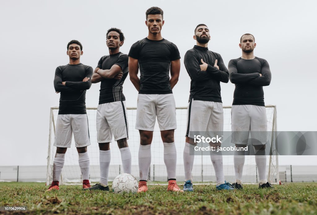 Footballers standing side by side on a soccer field Soccer players standing together with arms crossed on a soccer field. Low angle view of football players standing on soccer field during practice. Soccer Stock Photo