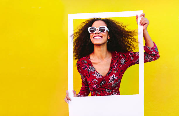 Beautiful female with big picture frame Mixed race woman with curly hair wearing sundress and sunglasses looking through blank photo frame. Beautiful female with big picture frame against yellow background. sunglasses photos stock pictures, royalty-free photos & images