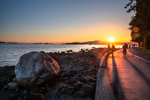 People on the Seawall around Stanley Park in Vancouver, Canada, at Sunset. Some Ships Anchored in English Bay are Visible in Background. Lens Flare.