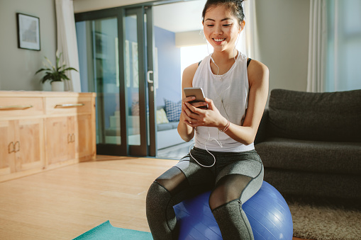 Woman listening to the music while sitting on a exercise ball at home. Happy sporty girl with smartphone relaxing on a fitness ball.