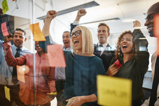 Businesspeople cheering while brainstorming with sticky notes in an office Diverse group of businesspeople cheering and fist pumping while brainstorming together with sticky notes on a glass wall in an office cheering stock pictures, royalty-free photos & images