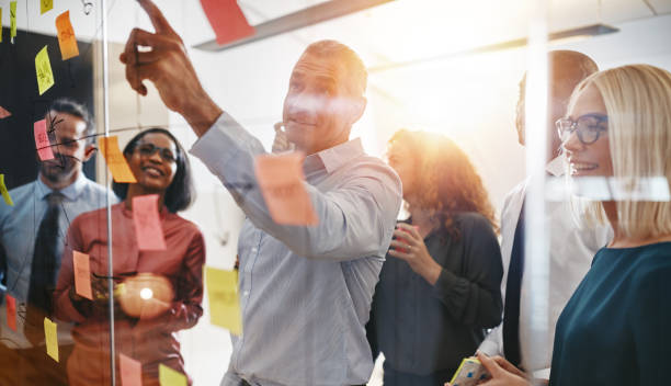 Businesspeople brainstorming together with sticky notes in an office Smiling mature manager pointing at sticky notes on a glass wall while brainstorming with his diverse team in a modern office brainstorming stock pictures, royalty-free photos & images