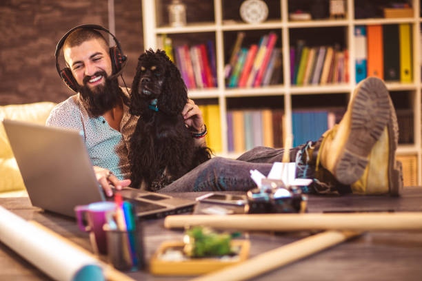 bell'uomo tatuato che lavora a casa su laptop mentre è seduto al tavolo con un cane carino - working at home headset telecommuting computer foto e immagini stock