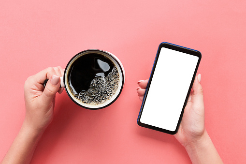 Female hands holding black mobile phone with blank white screen and mug of coffee. Mockup image with copy space. Top view on pink background, flat lay.