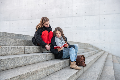 Two young  woman sitting on stone stairs outdoors reading together in booklet