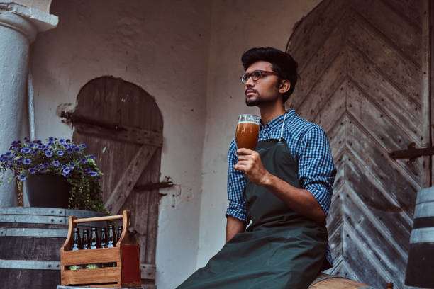 maître-brasseur, assis sur un tonneau en bois et tient un verre de bière artisanale, se détend après le travail. - after work beautiful people beer beer bottle photos et images de collection