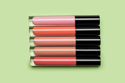 Five brightly coloured bottles of liquid lipstick and lip gloss arranged in a row on clean pastel green background shot with studio light from above