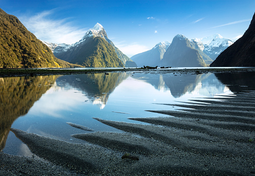 Ripples in the sand point to the tall peaks of Milford Sound which are reflected in the still waters of the fiord.\nMilford Sound is situated in Fiordland National Park, on the south west of New Zealand's South Island. It is known for its shear cliffs rising straight out of the sea. Mitre Peak (centre left) is a famous landmark and rises 5,560 feet (1,690m) from sea level.