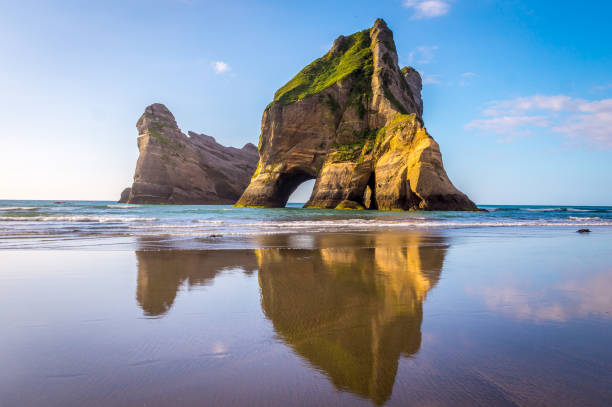 Wharariki Beach, New Zealand: Beautiful rock formations reflect in the water as mighty waves clash against the shore. This vast stretch of coast displays natural beauty in so many ways.