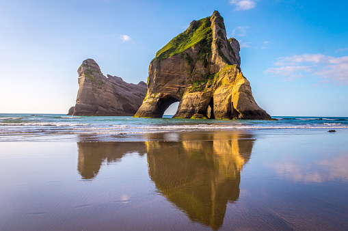 Wharariki Beach, New Zealand: Beautiful rock formations reflect in the water as mighty waves clash against the shore. This vast stretch of coast displays natural beauty in so many ways.