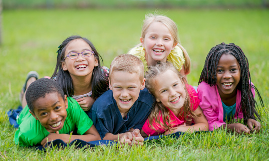 A happy, diverse group of primary school age children lie together in the grass in the park.