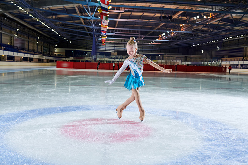Full length portrait of talented little girl figure skating in indoor rink during performance in spotlight, copy space