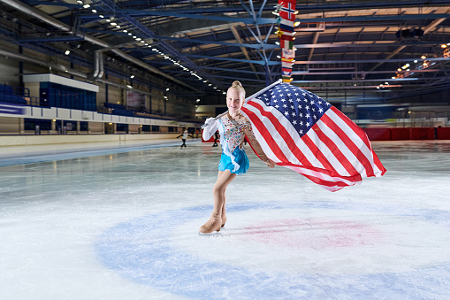 Full length portrait of smiling little girl figure skating carrying American flag in spotlight and looking at camera, copy space