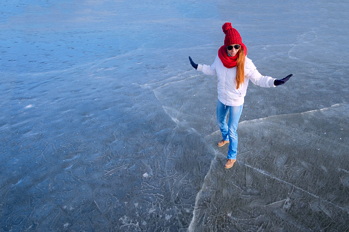 Girl in sunglasse, white jacket, red cap and red scarf holding balance and having fun while walking on ice on the frozen lake in winter