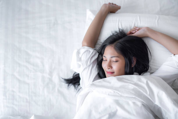 Beautiful young pretty Asian woman wake up and make happy smile with white shirt at the white bed in the morning. stock photo