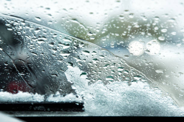 Snow-covered car windshield with melted snow drops and windshield wipers. Blurred lights of passing cars on the background. Copy space.