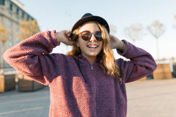 ritratto all'aperto di una ragazza adolescente sorridente di 13, 14 anni con cappello e occhiali da sole. sfondo città, ora d'oro. - teenager 14 15 years 13 14 years cheerful foto e immagini stock