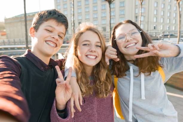 retrato de tres amigos adolescentes niño y dos niñas sonriendo y tomando un selfie al aire libre. hora de fondo, oro de la ciudad - three boys fotografías e imágenes de stock