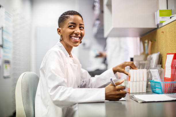 Smiling chemist working at desk Female chemist sitting at her desk and looking away smiling. African woman pharmacist working at her desk. laboratory stock pictures, royalty-free photos & images