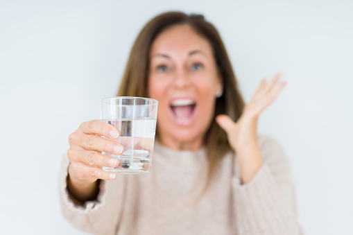 Middle age woman drinking glass of water isolated background very happy and excited, winner expression celebrating victory screaming with big smile and raised hands