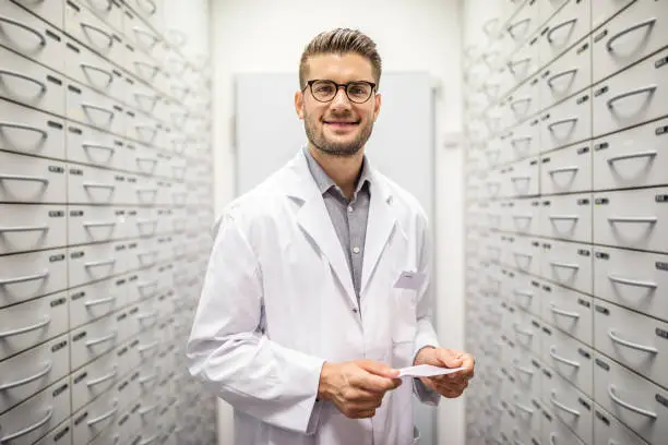 Portrait of a young male pharmacist holding medicine prescription standing in storage room in pharmacy.