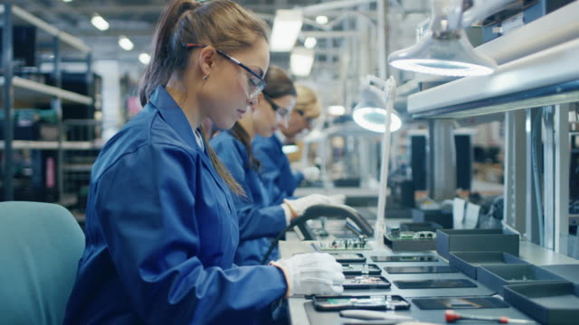 Female Electronics Factory Worker in Blue Work Coat and Protective Glasses is Assembling Printed Circuit Boards for Smartphones with Tweezers. High Tech Factory with more Employees in the Background.