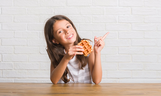 Young hispanic kid sitting on the table eating waffle very happy pointing with hand and finger to the side