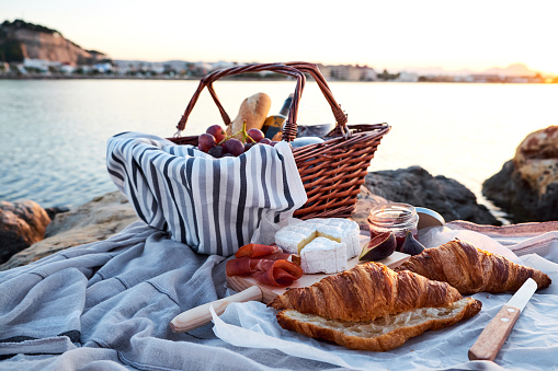 Beautiful romantic picnic composition with basket, croissants, red wine, bread, jam, cheese and jamon on the beach close to the sea.