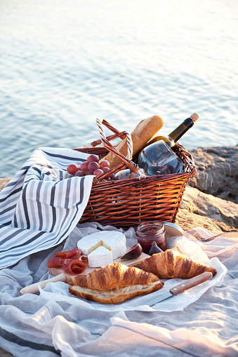 Picnic basket with red wine, bread, jam, cheese, croissant and jamon on a sunny day. Romantic picnic on the beach with sea on background.
