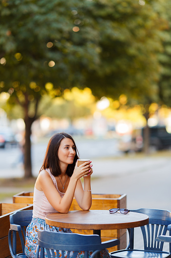 Portrait Of A Beautiful Smiling Happy Lady With A Hot Coffee Sitting On a Summer Terrace In A Cafe. A Young Girl Is Sitting And Drinking Coffee At A Restaurant Table.