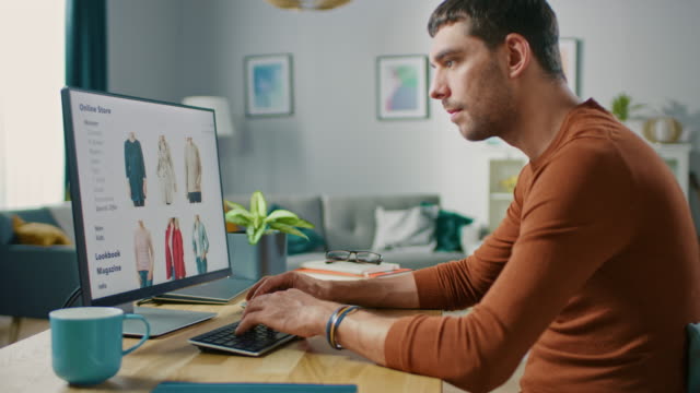 Handsome Young Man Sits at Home Browses Through Online Retailer Clothes Selling Store. Man Does Online Shopping for Clothing Items as a Gift for His Wife.