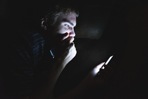 The image displays a teenager reading shocking news on his cell phone as he is lying on a couch in the dark.