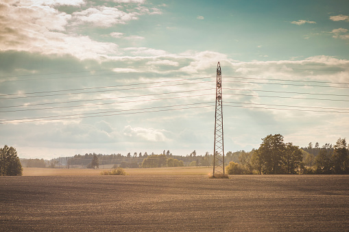 Dusty agricultural landscape with an electrical supply tower in a field with bushes littering the background. Clouds populate the cyan sky of an autumn day, after a long hot summer.