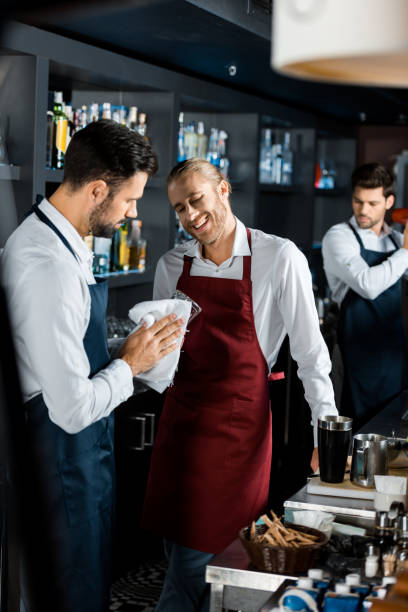 istock cheerful smiling barmen standing at workplace while coworker polishing glass 1071027320