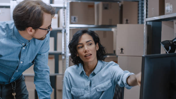 istock Male and Female Warehouse Inventory Managers Talking, Using Personal Computer and Checking Stock. Female Pointing at Screen. In the Background Rows of Shelves Full of Cardboard Box Packages. 1071028826