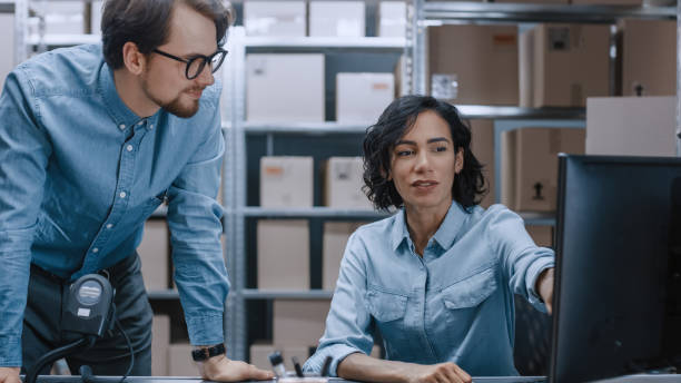 Male and Female Warehouse Inventory Managers Talking, Using Personal Computer and Checking Stock. Female Pointing at Screen. In the Background Rows of Shelves Full of Cardboard Box Packages. Male and Female Warehouse Inventory Managers Talking, Using Personal Computer and Checking Stock. Female Pointing at Screen. In the Background Rows of Shelves Full of Cardboard Box Packages. warehouse office stock pictures, royalty-free photos & images