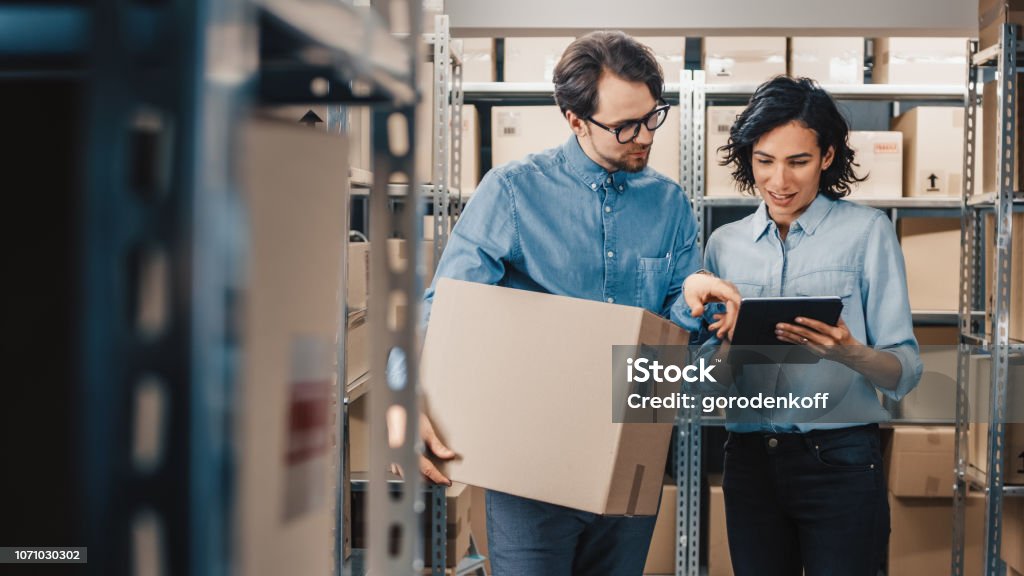 Female Inventory Manager Shows Digital Tablet Information to a Worker Holding Cardboard Box, They Talk and Do Work. In the Background Stock of Parcels with Products Ready for Shipment. Warehouse Stock Photo