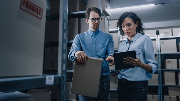 istock Female Inventory Manager Shows Digital Tablet Information to a Worker Holding Cardboard Box, They Talk and Do Work. In the Background Stock of Parcels with Products Ready for Shipment. 1071030426