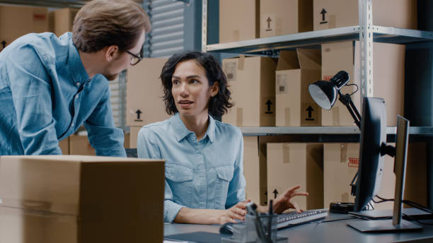 istock Male and Female Warehouse Inventory Managers Talking, Using Personal Computer and Checking Stock. In the Background Rows of Shelves Full of Cardboard Box Packages. 1071030500