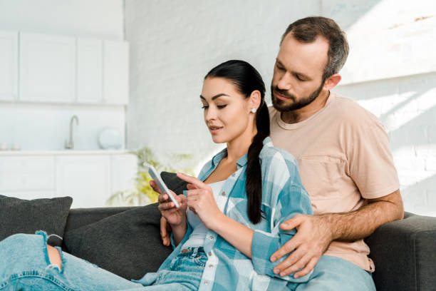 istock husband and wife sitting on couch and using smartphone at home 1071052170